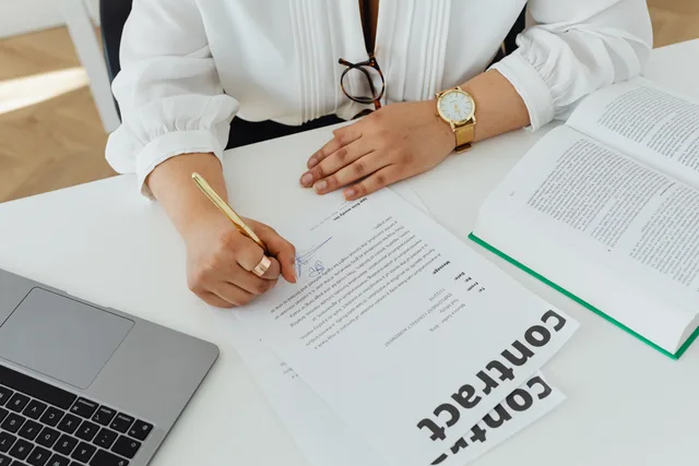 Close-up of a businesswoman signing a contract at her desk, with a laptop and book. Aftaler og compliance med underleverandører i Danmark